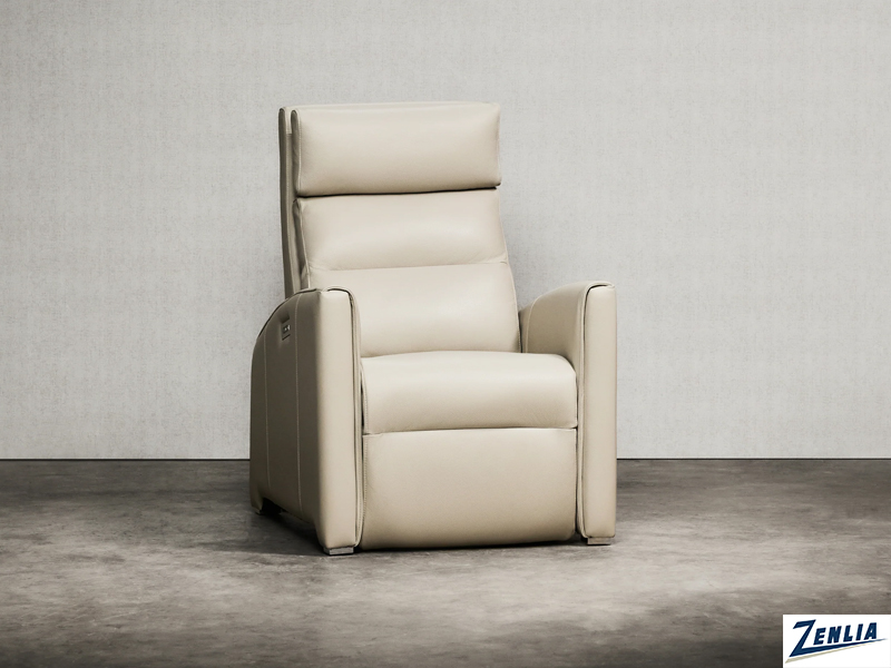 A sleek, modern recliner in light beige leather sits against a plain gray wall, featuring a tall backrest and cushioned armrests, with a small control panel visible on the side.