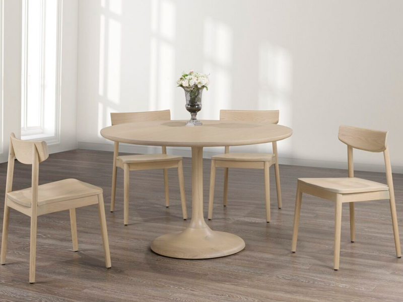 A round, light wood dining table with a pedestal base is surrounded by four matching chairs on a wooden floor. A small vase with white flowers is placed in the center of the table, and the room is softly lit by natural light from a nearby window.